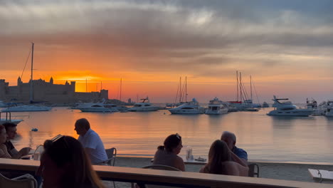 sunset view of a historic castle and marina with yachts and people dining outdoors
