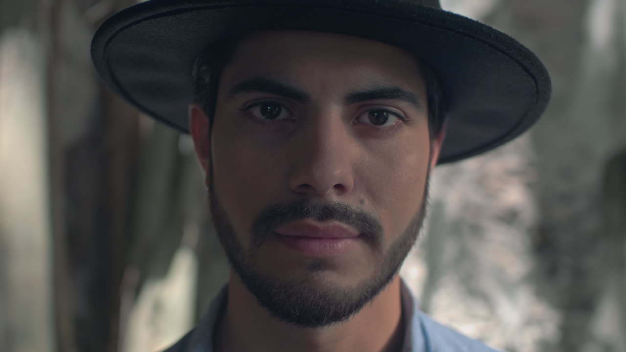 High-quality 4K footage of a stylish young man with a beard and hat, slowly opening his eyes in soft, natural light. He has a focused, mysterious expression. Ideal for travel, discovery