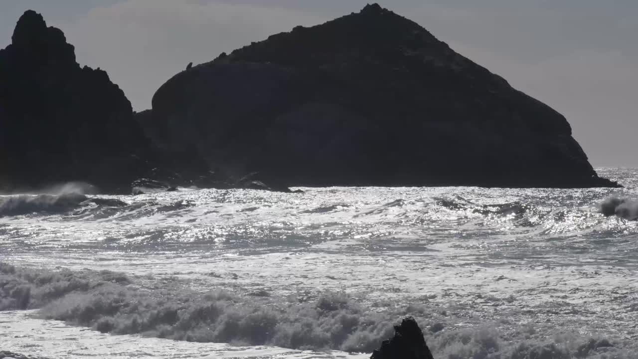Powerful Pacific Ocean waves shimmer in the sunlight and crash against dark sea stacks along the Big Sur coast.