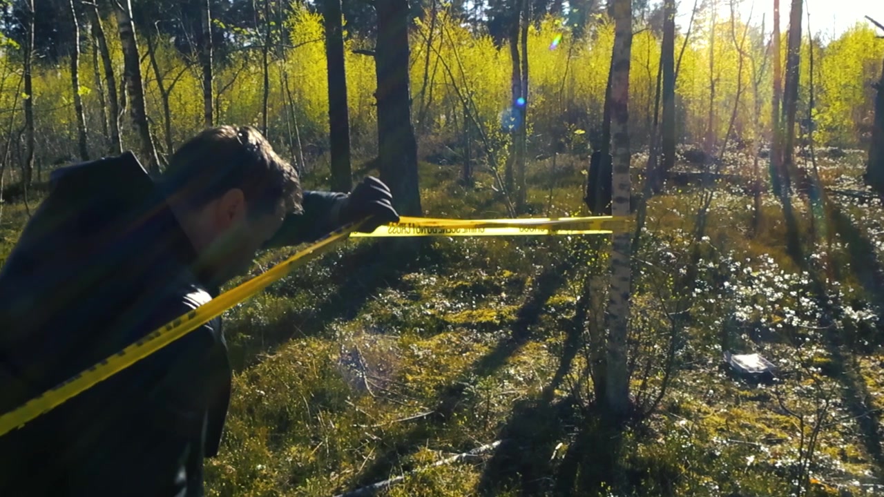 Mannlig politidetektiv i skinnjakke og hansker som går inn i et begrenset område under et gult sperrebånd for åsted i en solrik skog.