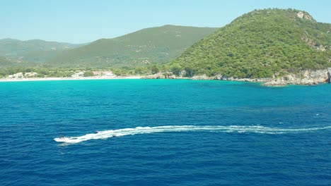 Drone-Sigue-A-Una-Lancha-Remolcando-Una-Balsa-Inflable-En-Aguas-Turquesas-En-La-Playa-De-Karavostasi,-Grecia