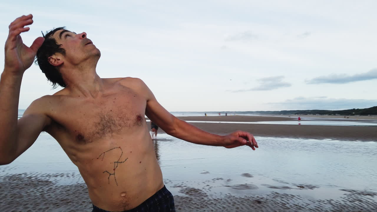 Barefoot man in swim trunks misses a frisbee across shallow tidewater on Omaha Beach, with Normandy’s green coastline and coastal homes in the background, captured in slow motion