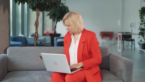 retrato de una mujer mayor con un traje de negocios rojo, sentada en un sofá en un centro de negocios, trabajando en su computadora portátil, recibe un mensaje alegre de victoria o éxito y comienza a regocijarse con una sonrisa