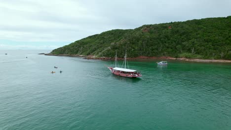 Aerial-orbit-of-a-red-tourist-boat-in-crystal-clear-waters-next-to-the-dense-mountains-of-Tartaruga-beach,-Búzios,-Brazil