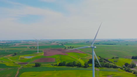 Renewable-energy-wind-turbines-rotating-above-flat-green-pastoral-farm-land-on-sunny-day,-rising-aerial