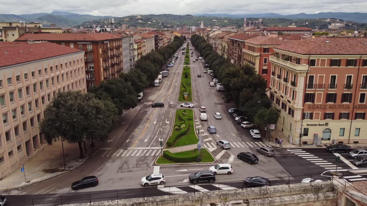 Static view of Viale del Piave, Verona, with cars filling both sides of boulevard wth manicured central median stretches toward the hills, while buildings and trees flank the bustling street scene.