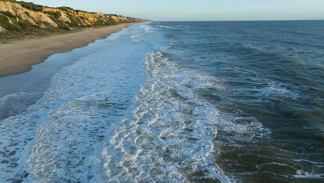 Aerial-view-tilting-over-waves-at-a-beach,-sunny-evening-on-the-coastline-of-Spain