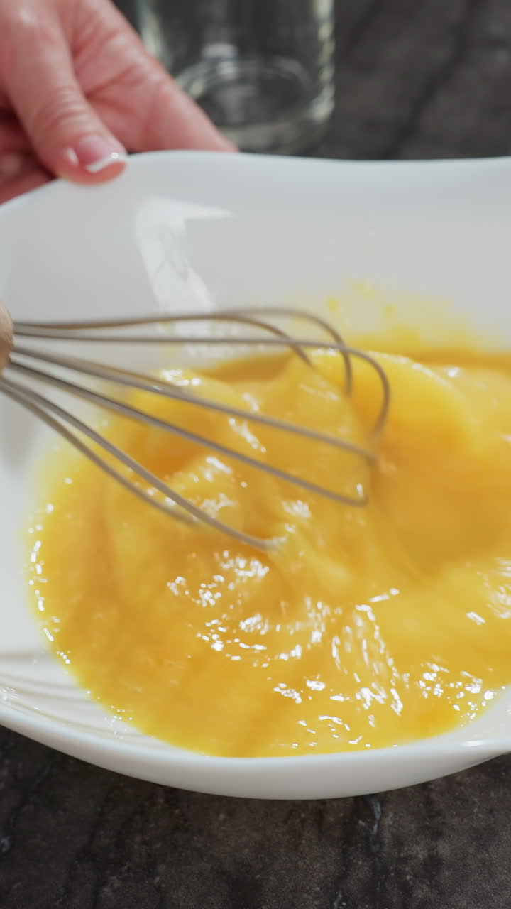 primer plano de un chef batiendo huevos en un cuenco blanco en una encimera oscura, la mano agita firmemente la mezcla, mezclando las yemas para hornear, el batidor de madera contrasta con el suave