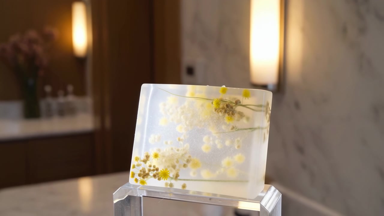 Bloc translucide décoratif contenant de petites fleurs séchées jaunes et blanches préservées, posé sur un piédestal transparent au premier plan d'une coiffeuse de salle de bain floue.
