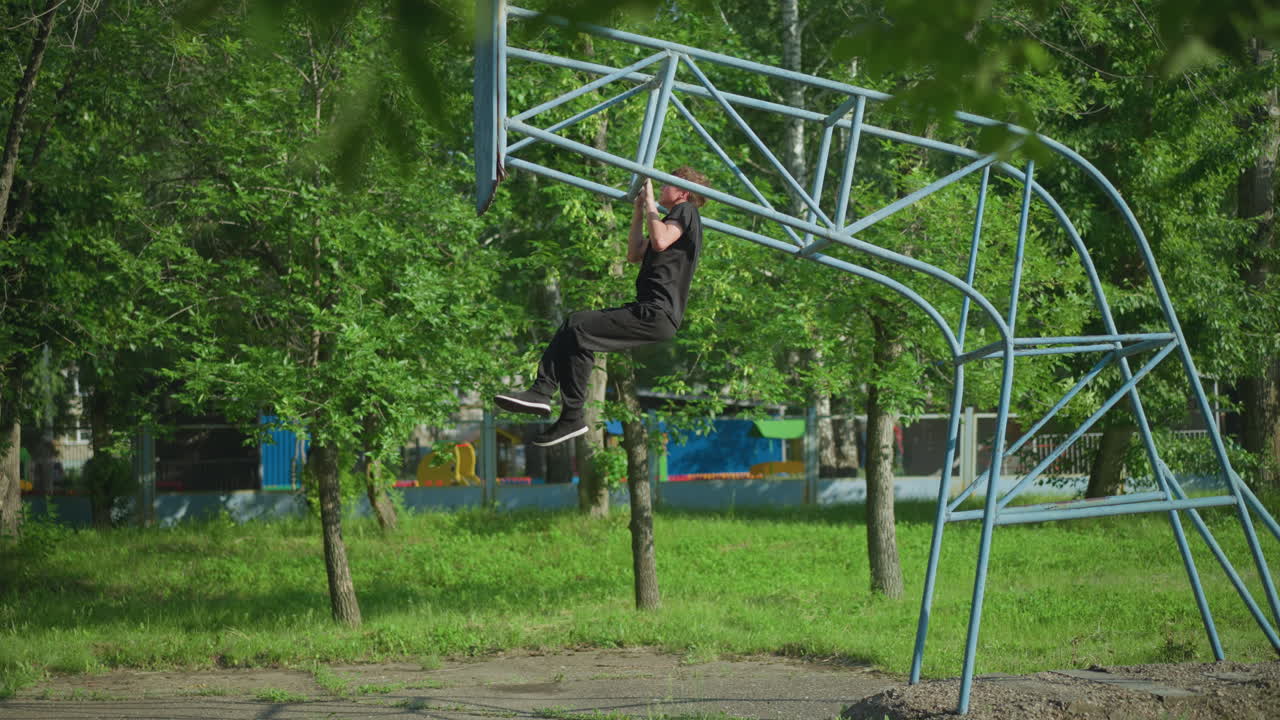 A young boy dressed in a black outfit is performing pull-ups on outdoor equipment, surrounded by a lush green environment with trees and a fence in the background