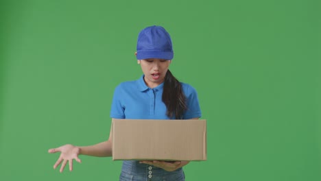 mensajera asiática en uniforme azul mostrando un gesto desconcertado y diciendo qué mientras entrega una caja de cartón en el fondo de la pantalla verde en el estudio