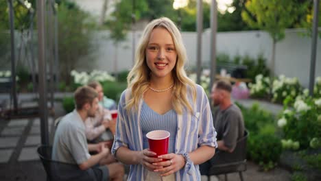 Portrait-of-a-happy-blonde-girl-in-a-blue-shirt-with-a-red-plastic-glass-in-her-hands-who-poses-and-smiles-while-relaxing-with-her-friends-in-the-courtyard-of-the-house