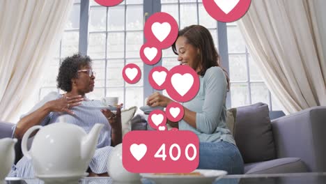 Animation-of-speech-bubble-with-numbers-and-heart-icons-over-two-women-talking-at-home