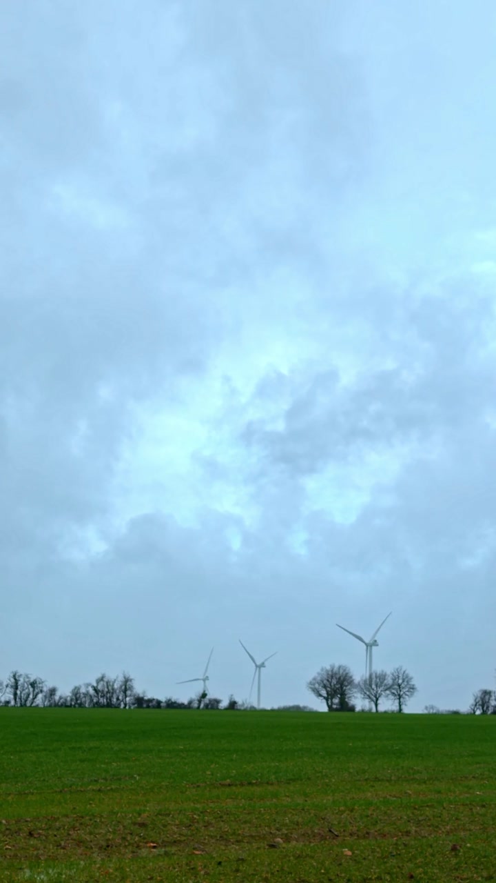 Vertical real-time shot of wind turbines spinning on hilltop horizon over green rural field under dramatic overcast sky in Latvian countryside