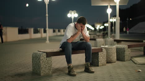 young man sitting on a bench outdoors at night, looking thoughtful and reflective, with city lights in the background, creating a calm and introspective atmosphere