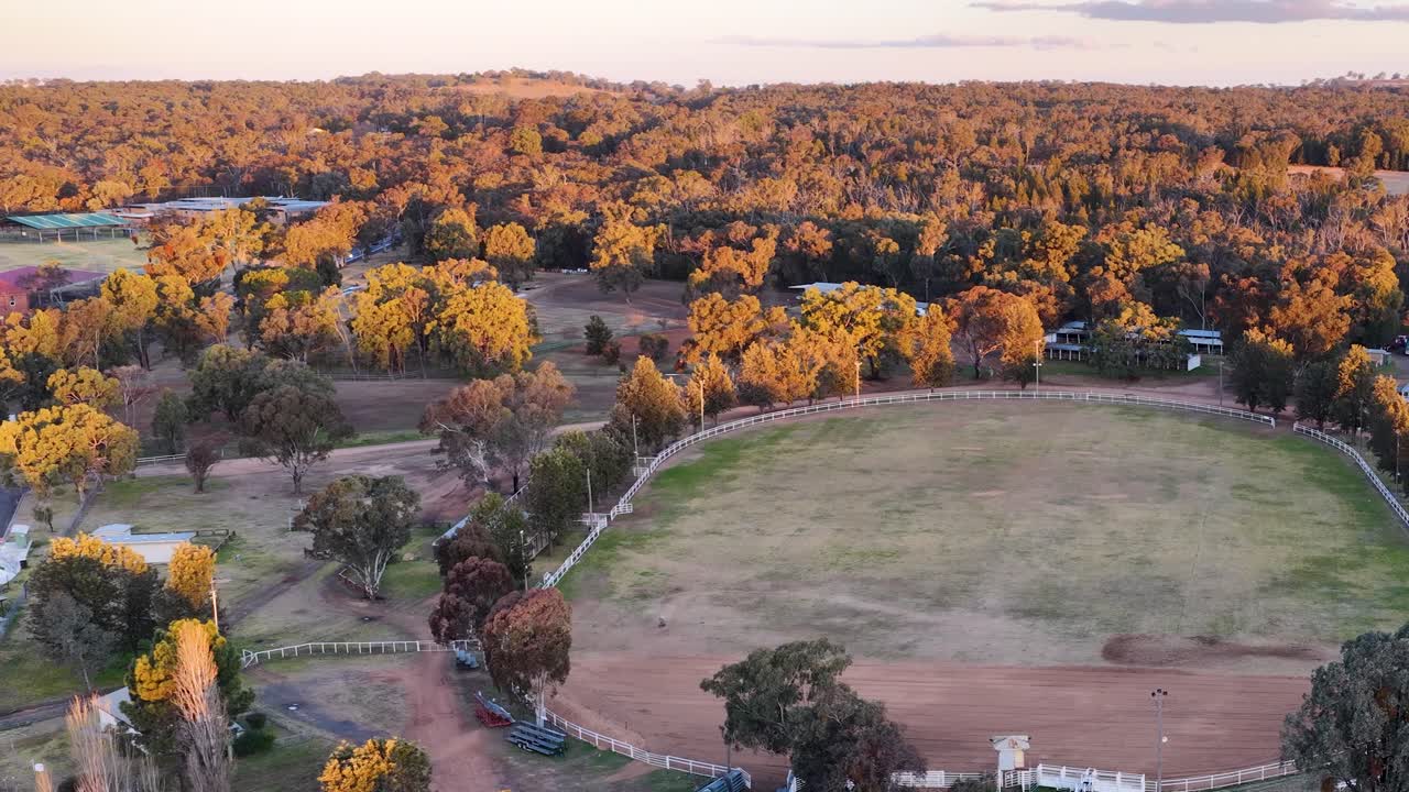 Aerial footage shows a drone smoothly flying over a rural football field surrounded by trees in golden sunset light, revealing nearby buildings and landscape