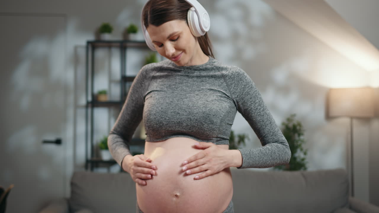 Femme enceinte portant des écouteurs et touchant son ventre tout en écoutant de la musique à l’intérieur