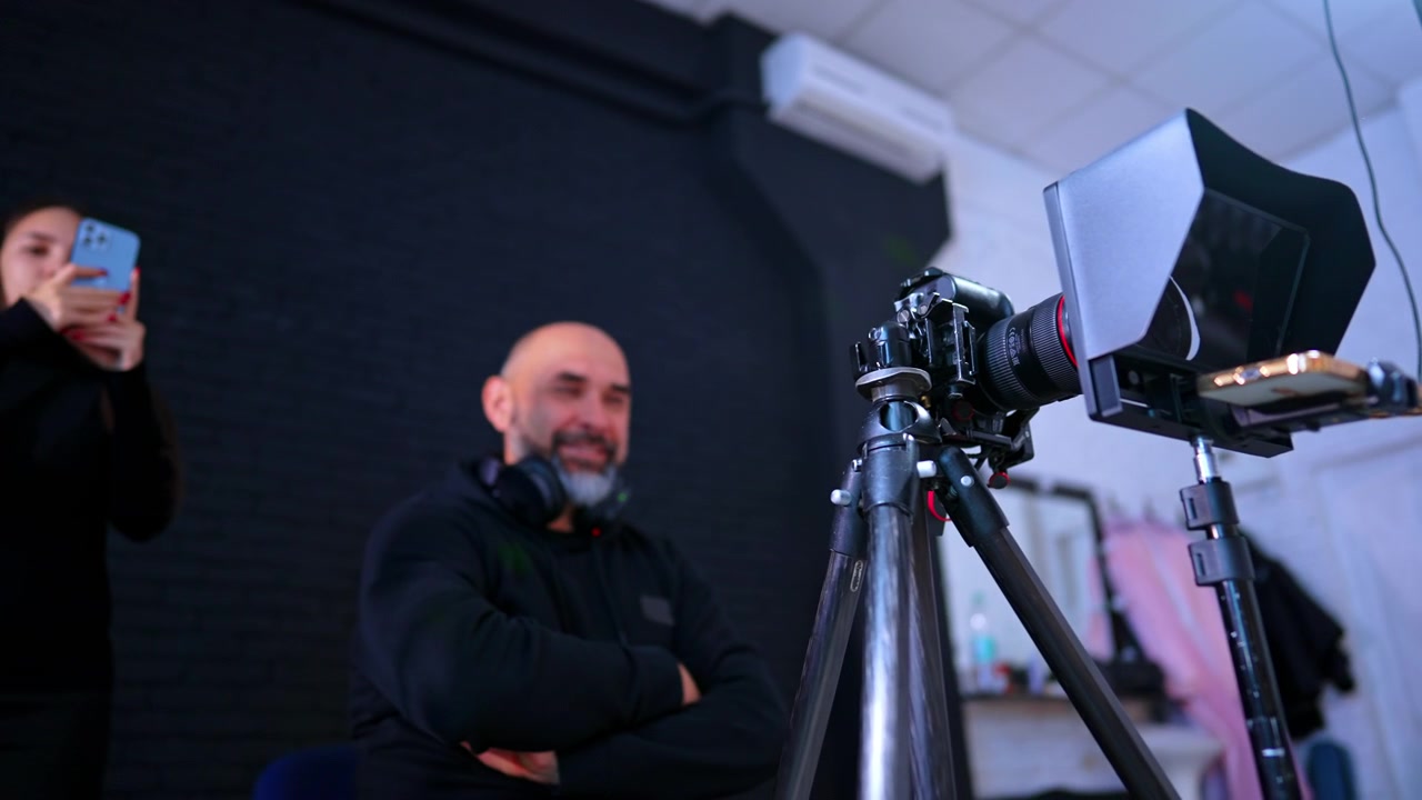 Professionelt kameraudstyr med teleprompter og mand i studie. Nærbillede af videokamera på stativ med teleprompterhætte i professionelt studiemiljø med mennesker.