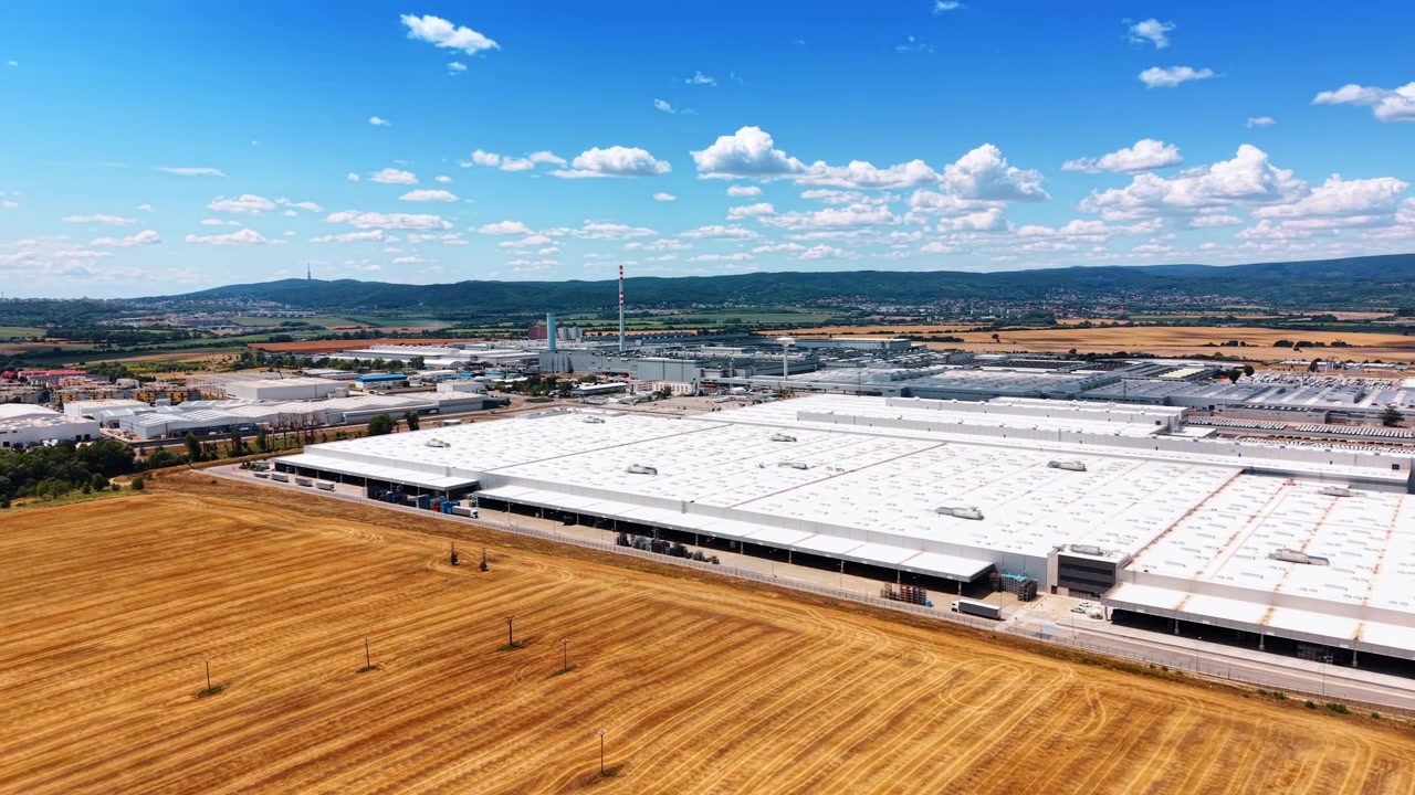 Complesso Industriale oltre un Campo Mietuto Dorato. Ampi Tetti Bianchi, Baie di Carico, Ciminiere, Strade e Distanti Colline Boscose Appaiono Sotto Nuvole Sparse