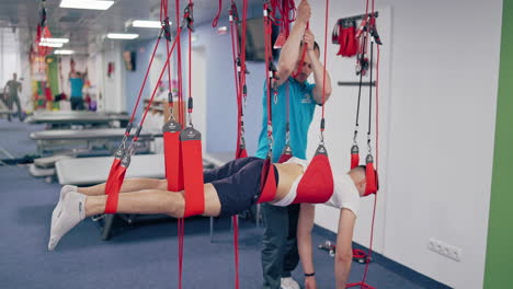 Instructor regulates the suspension rope system on the male in rehab center. Young man is hanging on special ropes to relieve back pain in a modern rehabilitation clinic.
