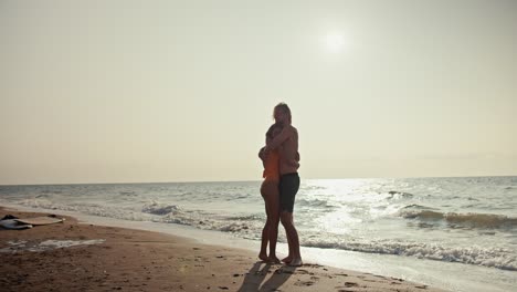 Happy-couple,-a-blond-guy-in-shorts-and-a-blonde-girl-in-an-orange-swimsuit-are-hugging-while-standing-on-a-brown-rocky-seashore-in-the-morning