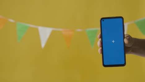 teléfono móvil con pantalla azul contra banderines en colores de la bandera nacional irlandesa colgado en el estudio para celebrar el día 1 de san patricio