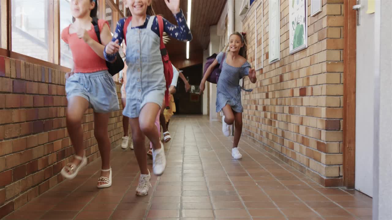 alumnas felices y diversas con bolsas escolares corriendo en el corredor de la escuela primaria