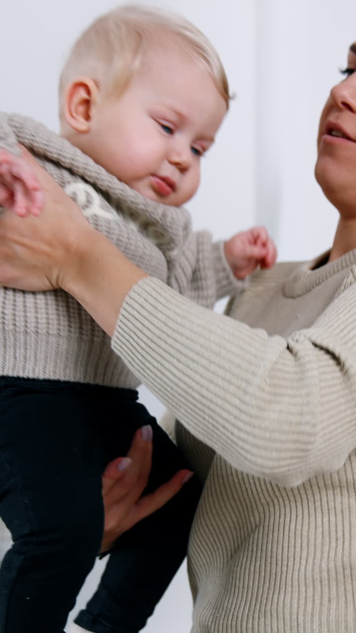 Adorable smiling blond baby in mom's hands. Mother and son wearing grey sweaters. Woman raises her child above head. Vertical video