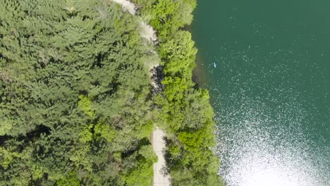 Vista-De-Pájaro-De-Un-Todoterreno-Que-Transporta-Dos-Motos-De-Agua-Detrás-De-La-Conducción-En-Una-Estrecha-Carretera-Cubierta-De-Bosque-Junto-A-Un-Hermoso-Lago-De-Agua-Verde-En-Wisconsin,-Ee.uu.