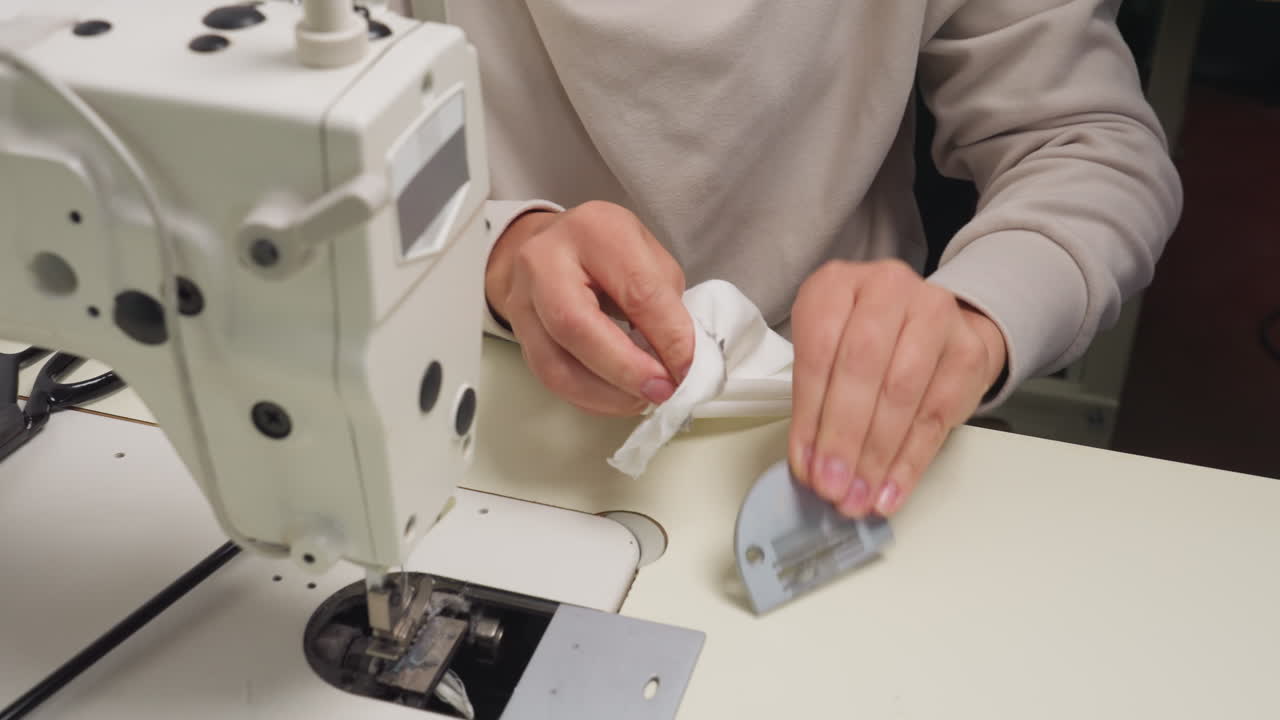 Designer cleaning slide plate of sewing machine using cloth for maintenance, table holds electric machine and scissors as person focuses on fabric hygiene, and machine care being practiced
