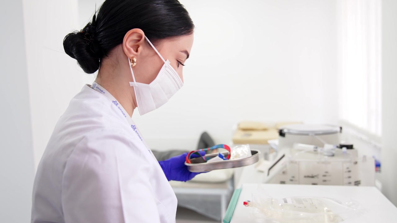 Infermiera caucasica con i capelli castani tiene un vassoio con il materiale per il prelievo del sangue. La sanitaria si avvicina al paziente uomo sdraiato sul lettino sullo sfondo
