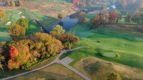 Campo-De-Golf-En-Otoño,-Trabajador-En-Fertilizantes-De-Tractor-Hermoso-Campo,-Puente-Que-Cruza-El-Río,-Toma-Aérea-En-órbita