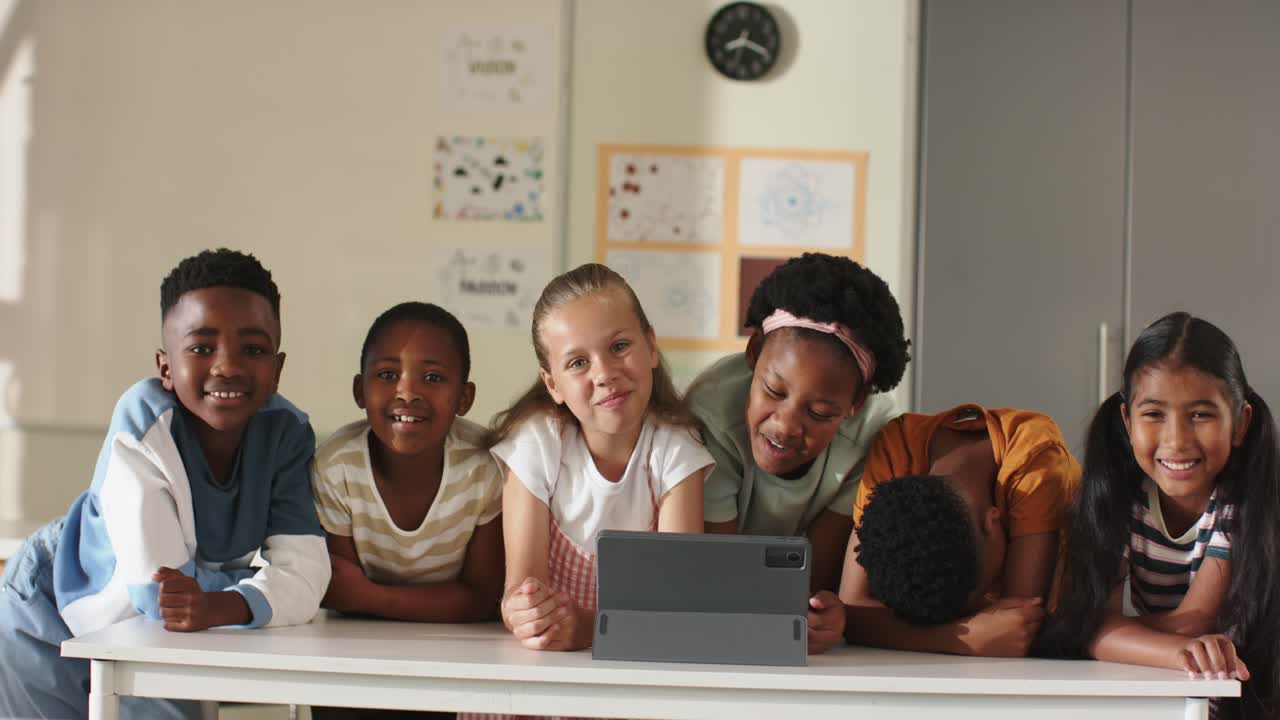 Schoolchildren friends gathering around tablet in classroom, watching funny clip, laughing together