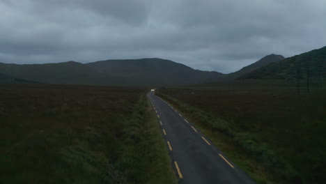 Forwards-fly-above-narrow-country-and-passing-against-car.-Cloudy-day-at-dusk.-Mountain-ridge-in-background.-Ireland