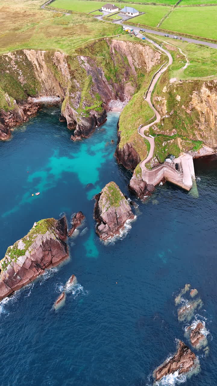 Some separate rocks stick out from water at the rocky shore of Ireland. Approaching the wavy tourist road descending to the water by the high mountain. Vertical video.