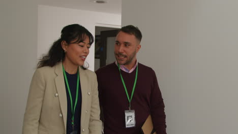 Male-And-Female-Teachers-Wearing-Security-Lanyards-Talking-As-They-Walk-Along-Corridor-In-School-Building-Together