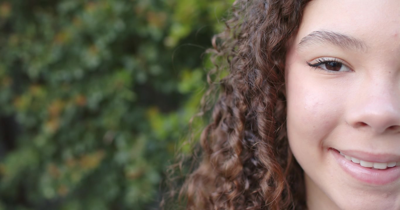 Adolescente afro-americana respondendo à câmera, piscando e sorrindo em uma cerca viva verde.