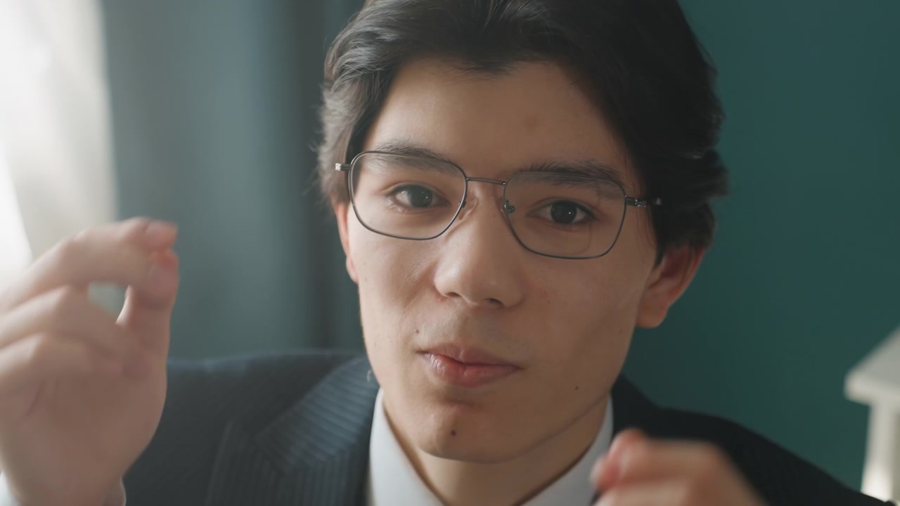 スーツ姿の若い男性が眼鏡を調整している。会議の準備中で、レンズの透明度を確認する様子。クローズアップのポートレートには思慮深い表情が浮かび、室内のオフィスを背景に柔らかな窓の光が差し込む。整頓された机と、そこはかとない緊張感が漂う。