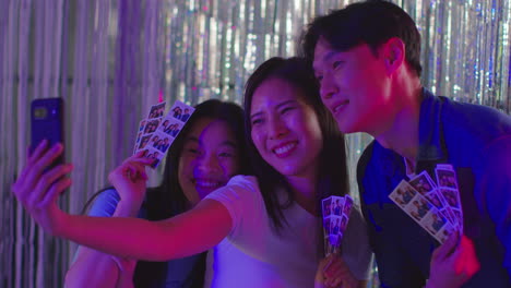 Group-Of-Friends-Taking-Selfie-Having-Fun-Looking-At-Prints-From-Photo-Booth-In-Nightclub-Or-Funfair-2