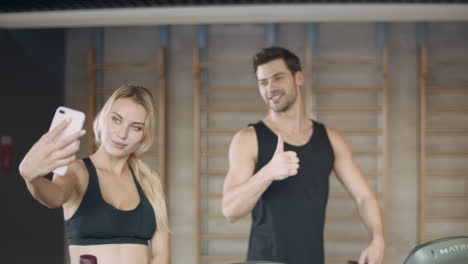 Serious-woman-posing-mobile-photo-at-fitness-training.-Man-making-yes-gesture