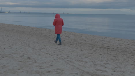 Person-Mit-Orangefarbenem-Regenmantel,-Die-An-Bewölkten-Tagen-Am-Sandstrand-Am-Meer-Spazieren-Geht