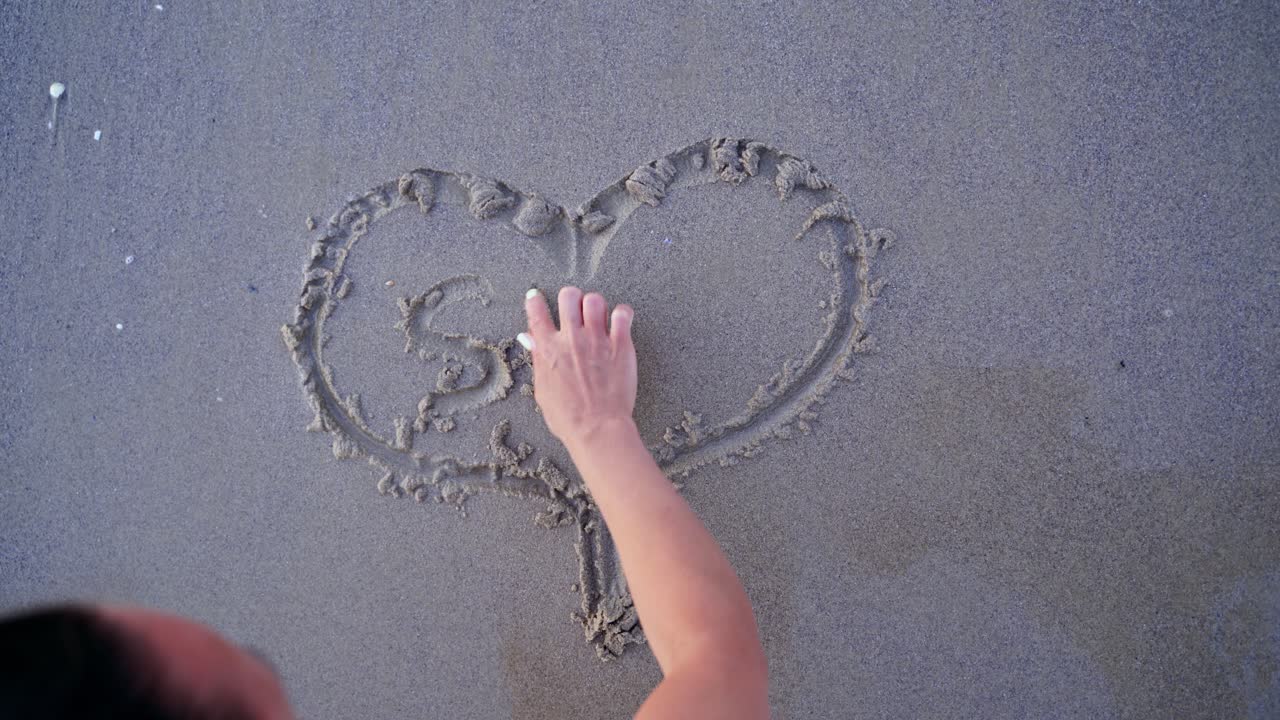 Woman draws heart on a wet sand. Female's hands writing the word sea on sand. Sea water washes away the drawing on a sandy beach.