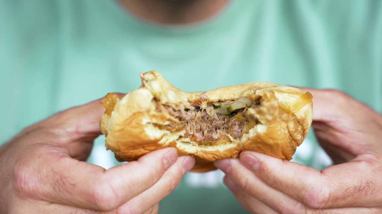 Slow motion close-up of a white man taking a big bite of a tasty smash burger, before revealing it to camera, as the cheese drips out next to his hands