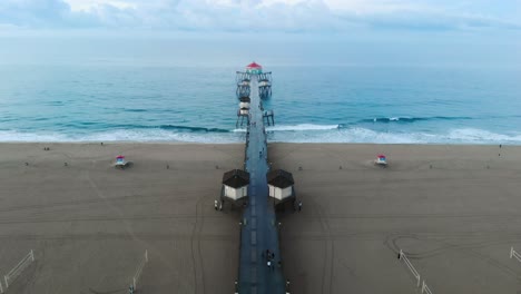 Eine-4K-Ansicht-Von-Ariel,-Die-Sich-Bei-Sonnenaufgang-Zum-Huntington-Beach-Pier-In-Der-Kalifornischen-Surf-City-USA-Bewegt,-Während-Surfer-Wellen-Schlagen-Und-Familien-Ihre-Reise-Und-Ihren-Urlaub-Am-Strand-Genießen