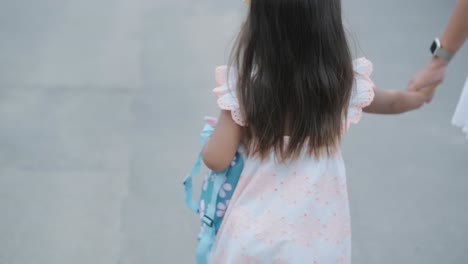 a pregnant woman and her young daughter, dressed in light summer outfits, walk hand-in-hand along a street in a residential neighborhood. the daughter holds a toy, enjoying the peaceful moment outdoor