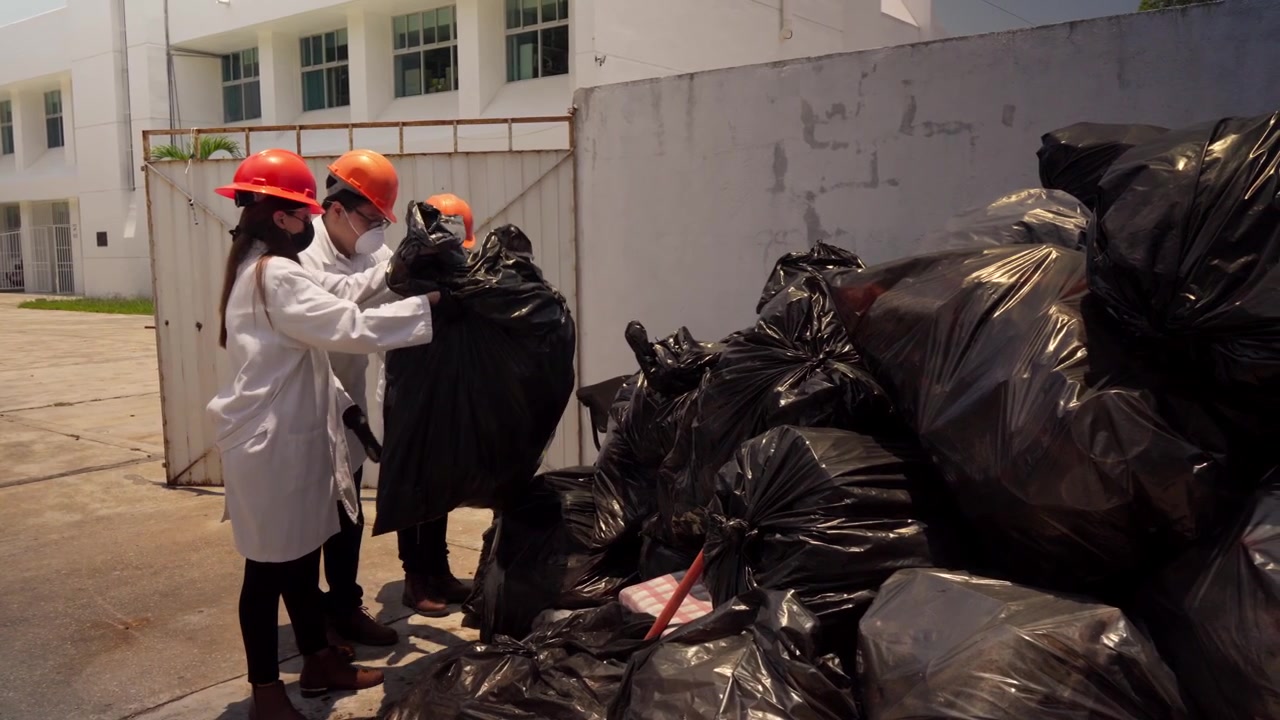 Tre lavoratori, due donne e un uomo, in un impianto di riciclaggio spostano sacchi neri della spazzatura per il processo di riciclo. Indossano camici bianchi da laboratorio e dispositivi di sicurezza, inclusi cappelli e mascherine