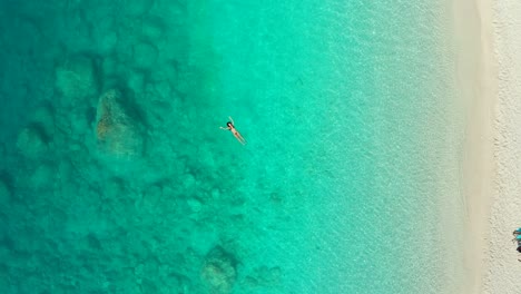 Vista-Aérea-Ascendente-De-Una-Joven-Nadando-En-Un-Mar-Turquesa-Transparente-En-Grecia,-Paisaje-Marino-De-Verano-Con-Una-Chica,-Playa,-Agua-Azul-Hermosa,-Vista-Superior-Desde-Un-Dron