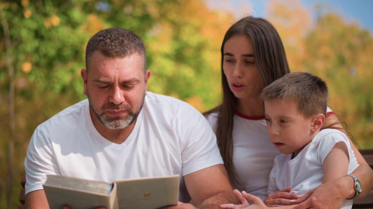 Primo piano di un uomo che legge un libro a un bambino, mentre una donna osserva, in un parco con alberi verdi e una panchina. L'uomo tiene in mano un libro rilegato aperto, il bambino è seduto sulle sue ginocchia e la famiglia interagisce insieme all'aperto.