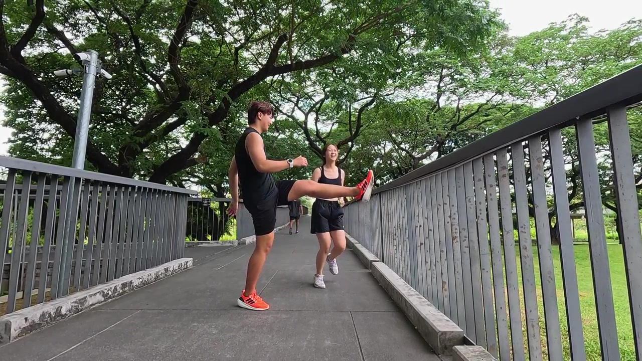 Pria dan wanita aktif mengenakan pakaian olahraga melakukan ayunan kaki di jembatan di bawah pepohonan rindang.