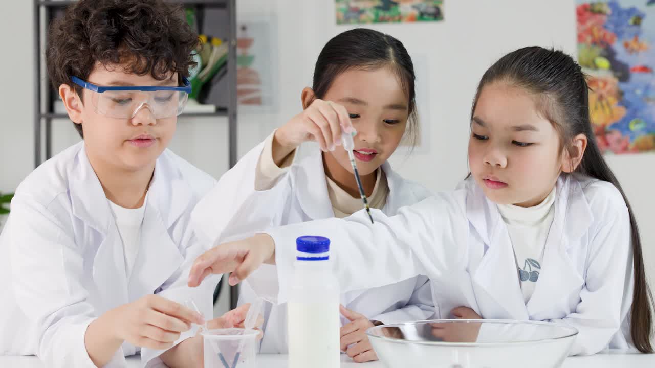 教室で、白衣を着たアジア人の子供3人が化学実験のためにピペットを使用している。